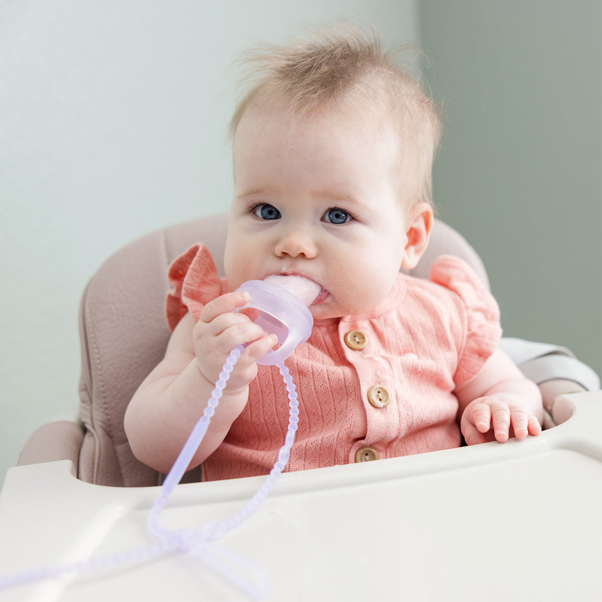 Infant in a highchair safely chewing on a pink Olababy silicone teether tether.