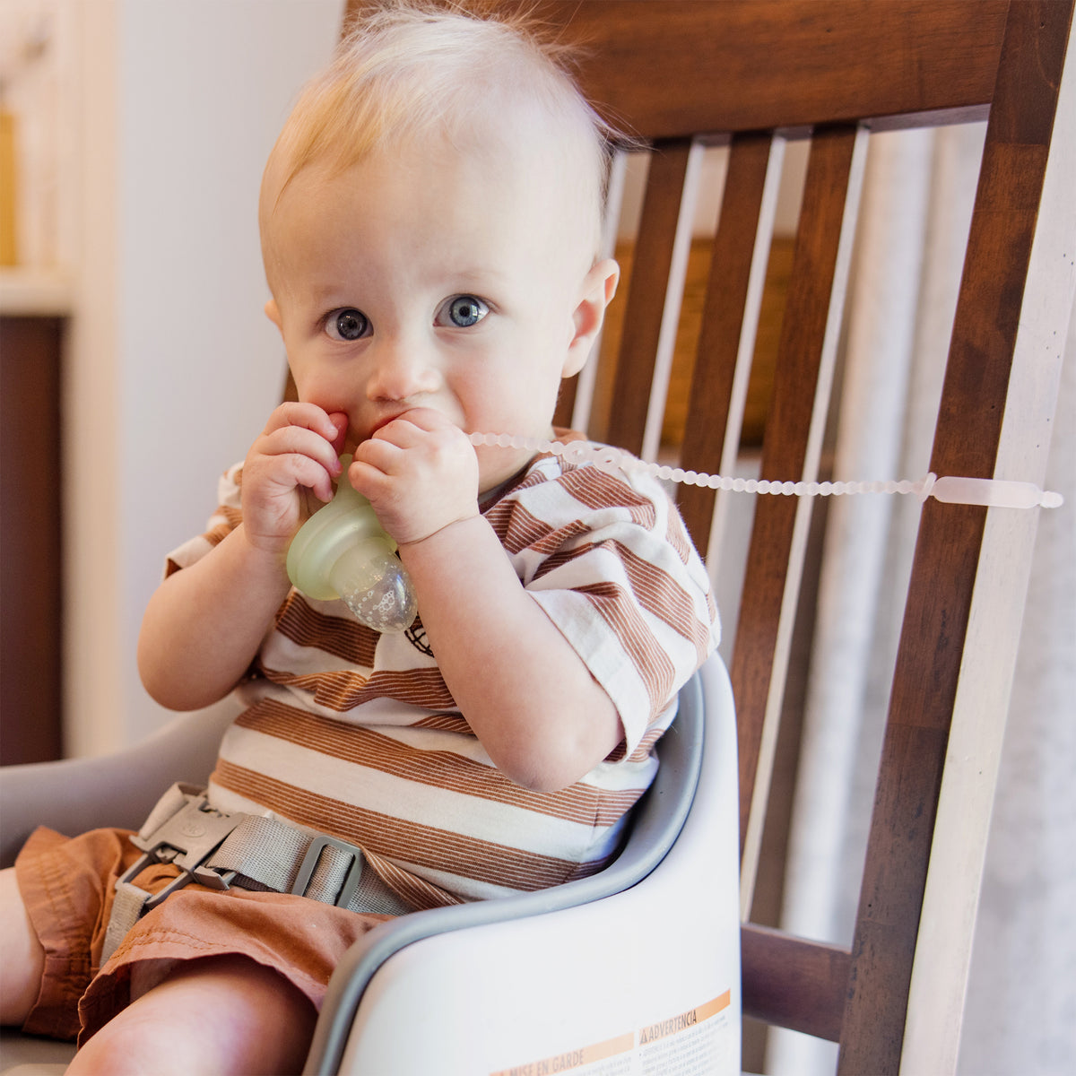 Baby holding a green silicone pacifier strap attached to a highchair to prevent dropping.